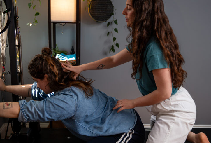 Sofia, a white brunette dressed in a blue shirt and white pants, reaches forward to frame with her fingers the cervical spine of a mover leaning forward in front of her. The wall behind them is grey with a bright lamp and leafy plants. Photo by Rachel Keane.