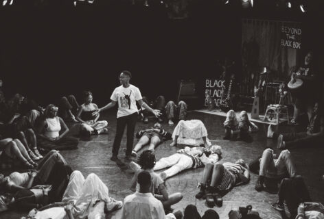 Many people lay and sit in a circle around one center person who is standing, guiding the group through community breath work. Photo by Kenny Rodriguez.