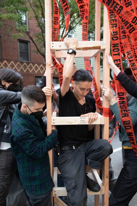 Castellanos is ascending a wooden structure that was built by the viewer's who are surrounding them, holding it steady. Castellanos has long black hair, wearing a black shirt and pants and has a raised arm holding themselves steady as they descend. Photo by Natalie Peart.