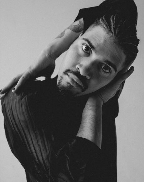 A black and white photo of Felix looking to the camera, in a vouge pose; their hands and forearms framing their face. Photo by Tamar.