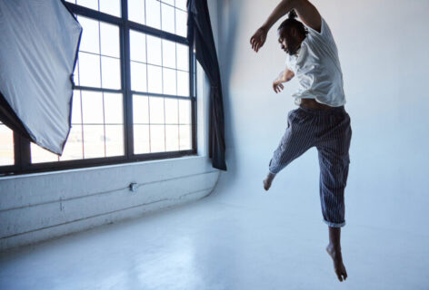 Jordan Demetrius Lloyd, a tall black man leaps in the air wearing black striped pants and a white shirt. His left leg is extended down toward the white floor while his right leg reaches toward the back corner of the room, which has a wall of windows with frosted glass. Photo by Whitney Browne.