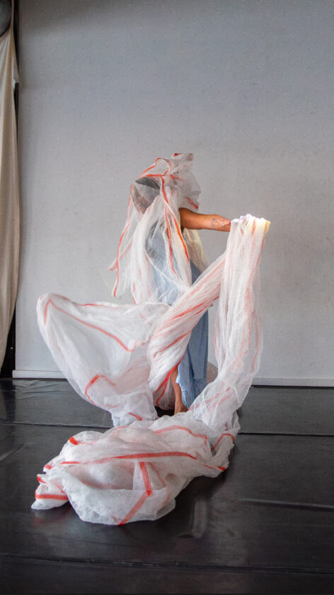 Nailah wears a blue dress. She tosses one arm to her side and one above her head while tangled in a red-trimmed, white mesh construction fabric.