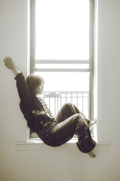 On a windowsill where soft lights are coming in, a person sits sideways, leaning back against one side with an arm stretched upward and legs bent toward the opposite wall. They have short hair with bangs and braids, wearing a loose dark shirt with wide black pants. Eyes are silently closed, dreaming.