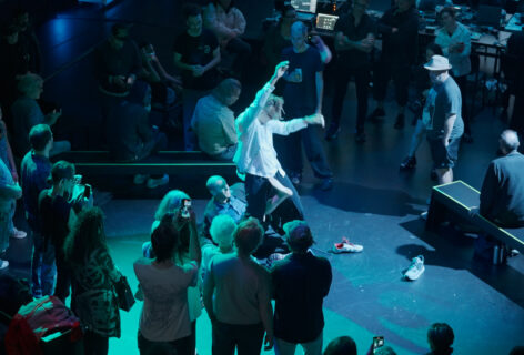 A man with dreadlocks in the center of a group of onlookers holds his arms up as he leans back and another man holds his feet. Photo courtesy the artist.