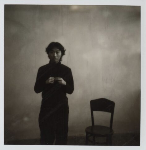 A black and white portrait photo taken when Yushu was acting in a short film. Dressed in all black, she stands to the left of a wooden chair with her hands held in front of her chest, touching her fingers together.