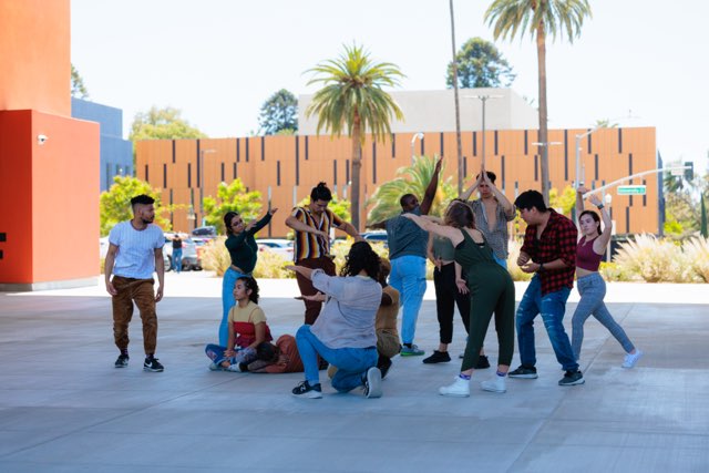 An image of many dancers outside standing on concrete. In the background a red-brown building and a palm tree can be seen. This is a group section from the piece titled “Stories Within” part of inVISIBLE/unHEARD April, 2022. The group performs at the Main Library breezeway, Riverside, CA. Photo by David Rodriguez. Pictured: Jessica Barajas, Aisha Shauntel Bardge, Gwen Benitez, Martin Carrillo, Al Ellison, Andy Garcia, Karla Sanchez, Norine Myers, Jonathan Ramirez, Edwin Antonio Sigüenza, Ernestina Soto, Chav’nair Stocker, Stephen Tanner