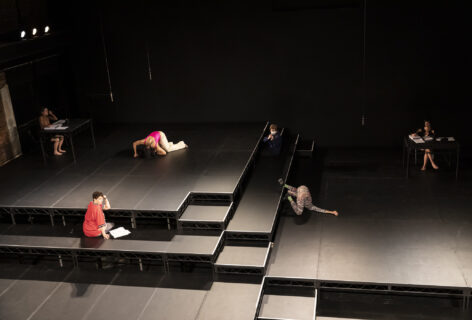 Remains performance photo; performers in various positions on a stage and risers, 2 sit at a table with papers in front of them. Photo by Rachel Papo.