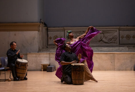 Milteri Tucker performing for Movement Research at the Judson Church. Milteri waves her large Bomba skirt. The skirt is purple and billows all around her. Two musicians play the drums. Photo by Rachel Keane.