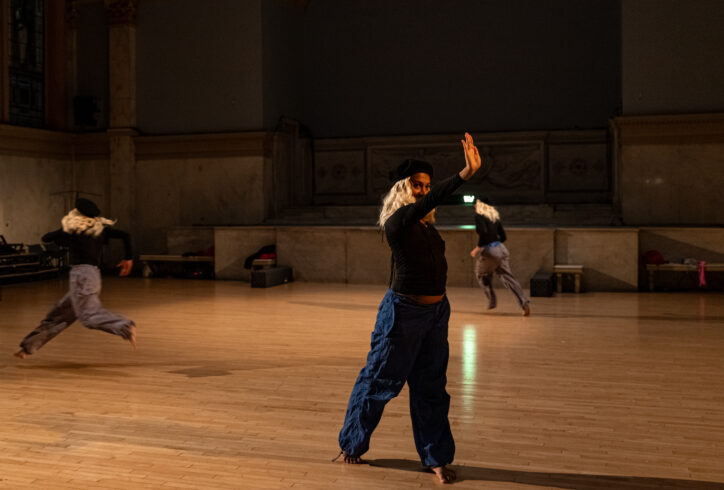 Sasha Vega's performance for Movement Research at the Judson Church. Three dancers wear blonde wigs and black berets. they move around the space. One dancer stands in the forefront a reaches. Photo by Rachel Keane.
