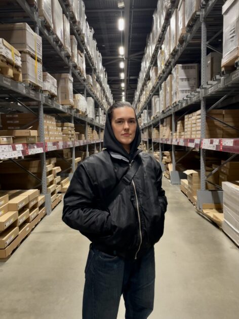 Molly, a white genderqueer person, stands in a black hoodie, bomber jacket and jeans in the walkway of a warehouse, looking at the camera. Behind them are long rows of shelves with cardboard boxes. Photo courtesy the artist.