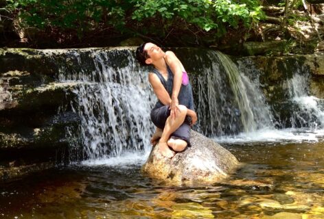 Water Spirit. A dancer perches on a large rock in golden-reflecting water. Their body seems to spiral as an extension of the rock, their face is turned upward towards the sunlight. A wide waterfall and bright green trees are behind them. Photo by Bea Ehrsam.