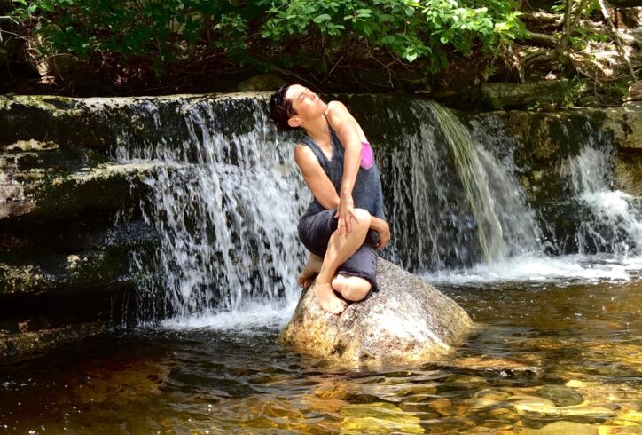 Water Spirit. A dancer perches on a large rock in golden-reflecting water. Their body seems to spiral as an extension of the rock, their face is turned upward towards the sunlight. A wide waterfall and bright green trees are behind them. Photo by Bea Ehrsam.