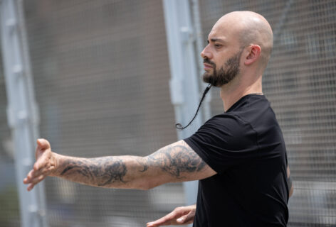 Christopher extends his right arm in the anterior-posterior plane. His long braided beard spirals around itself. His arms are heavily tattooed and he wears a black tee. Photo by Walter Wlodarczyk.