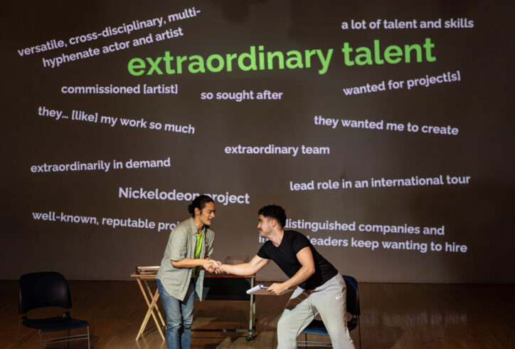 Production still of two performers shaking hands in front of a projected wall of English phrases. Photo by SD Herzog.