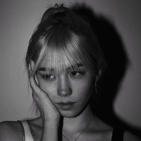 A young Asian woman with bleach-blonde hair, bangs, and a high ponytail sits in front of a plain background. She wears a tank top and a simple necklace. Her right hand is resting against her cheek as she looks off to the right of the camera.