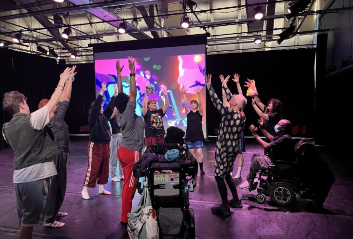 A group of people, some with wheelchairs, are reaching up, some with plush moths and tardigrades in their hands. They are growing a mushroom out of hyphae - in the background is the Planting Disabled Futures virtual reality world. Photo by Stephanie Heit.