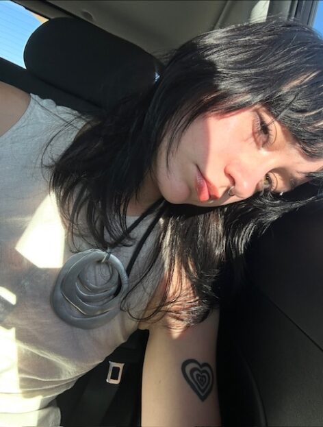 A young feminine person with dark hair and bangs sits in a car, wearing a necklace and nose ring, sunlight across their face, looking sideways.