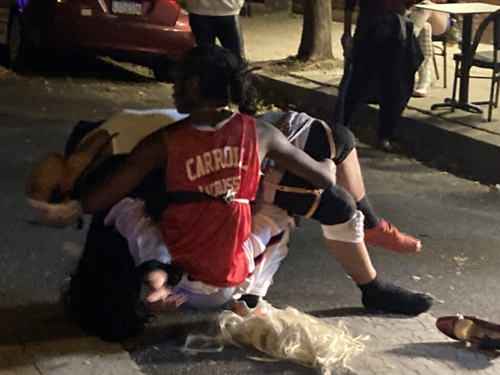 A still from Capeforce ii, a performance by Jordan Deal. Eden Lafayette, wearing red jersey, is kneeling on the ground holding Jordan Deal, whom is shown collapsed.