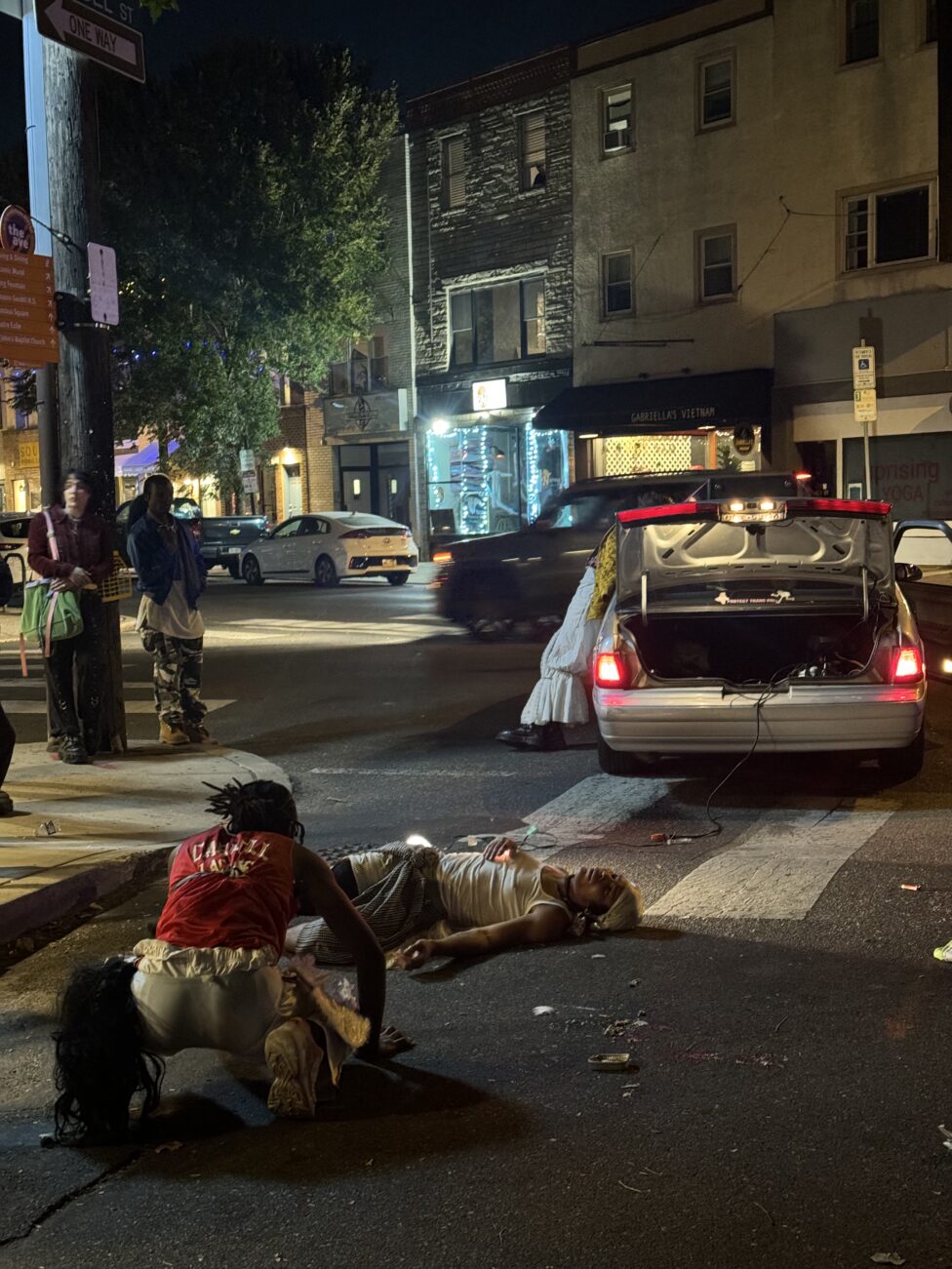 A still from Capeforce ii, a performance by Jordan Deal. Three performers (Jordan Deal, Eden Lafayette, and Vernon Jordan III) are shown collapsed and leaning across an intersection that has been blocked off by a car.